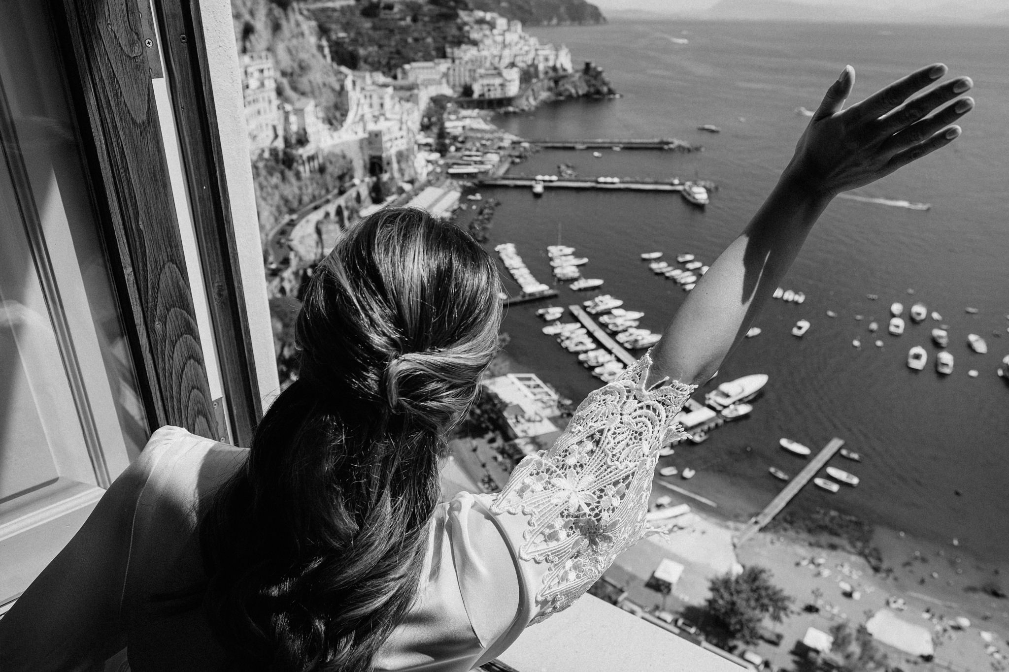 Bride waving from a high window over a harbour on the Amalfi Coast, boats and pastel houses far below