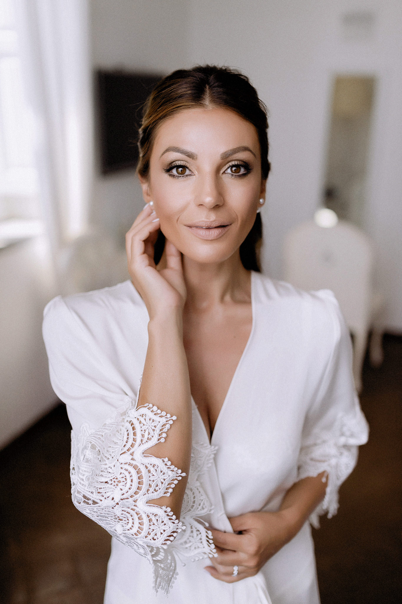 Bride in a white robe with scalloped lace sleeves, hair swept back, wearing pearl earrings