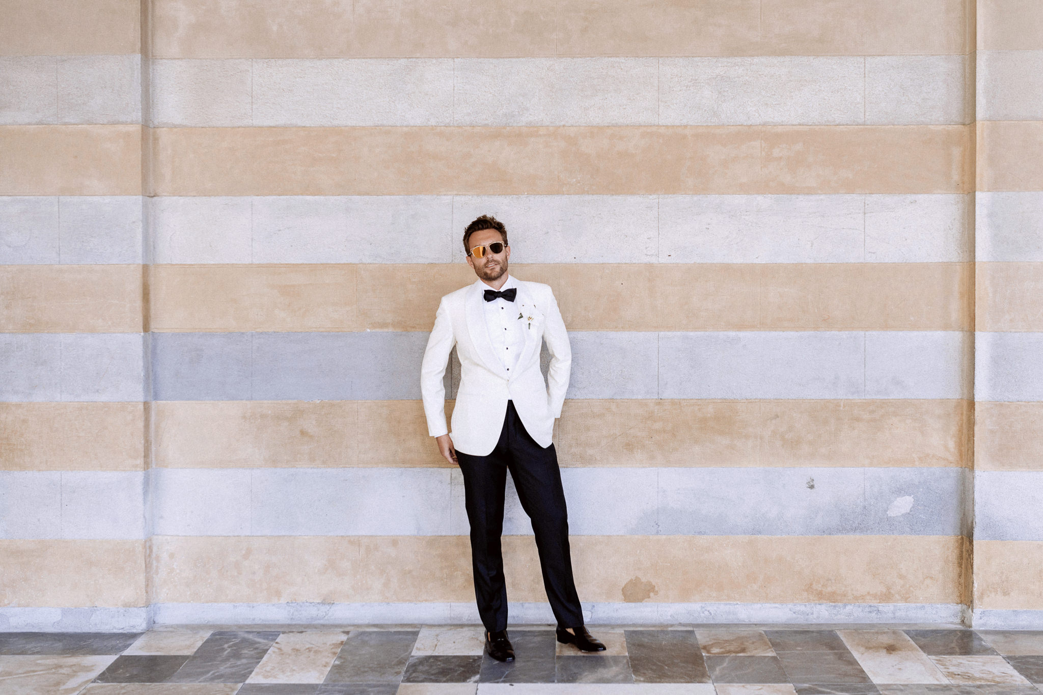 Groom in a white dinner jacket, black bow tie and sunglasses against a striped stone wall