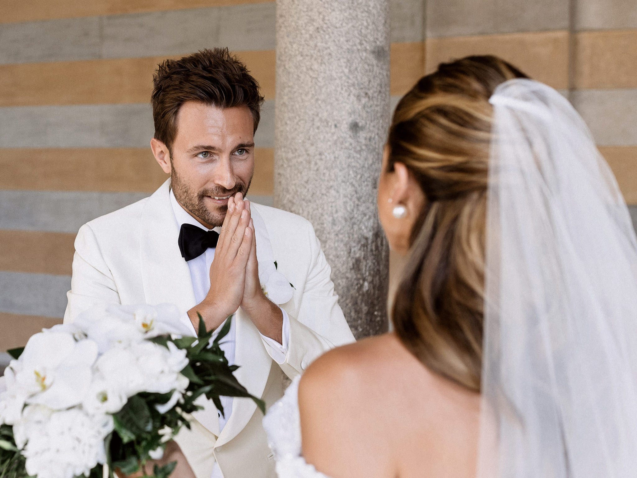 Groom pressing his hands together with emotion at the first look as the bride approaches with her orchid bouquet