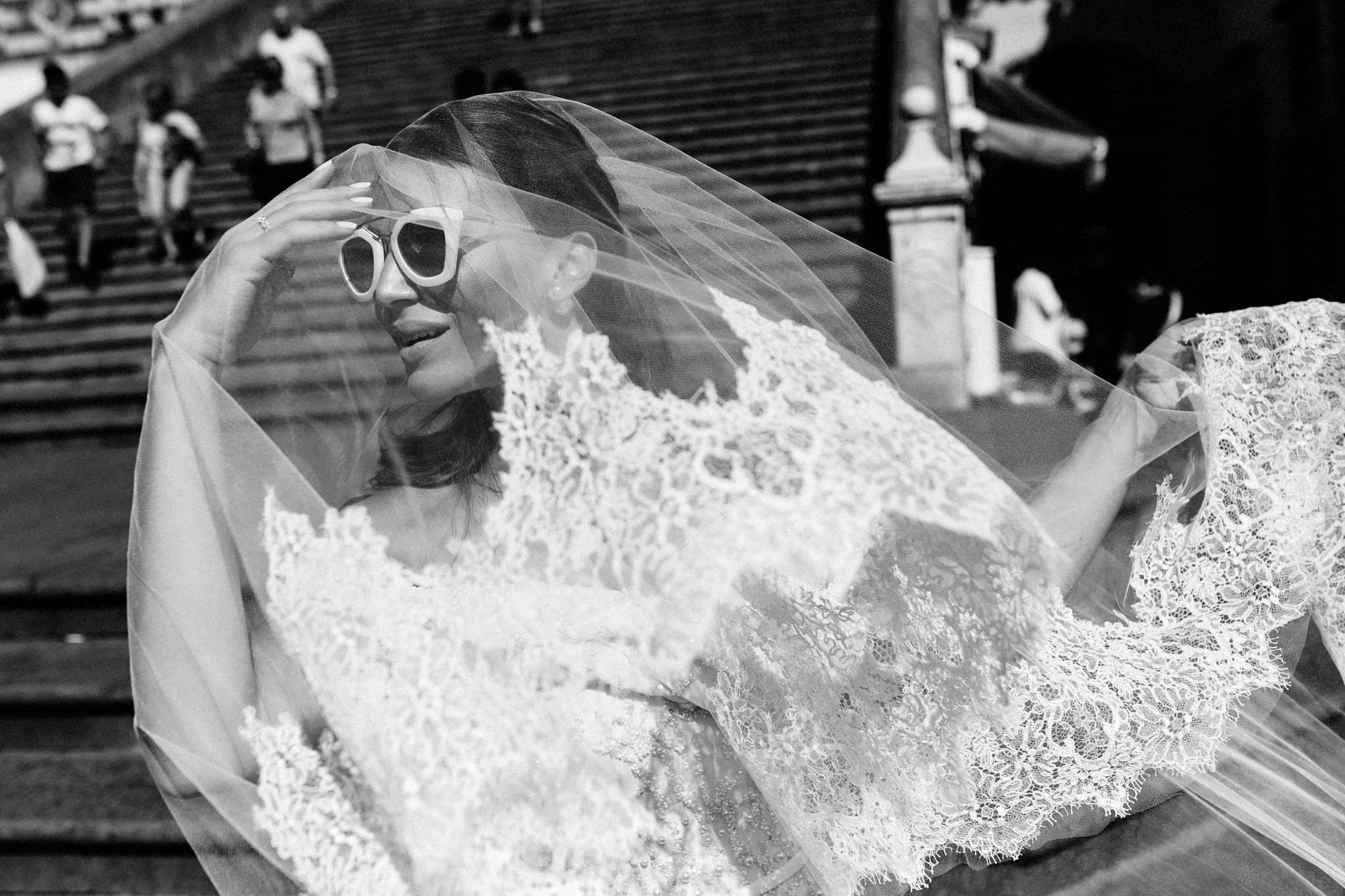 Bride in white sunglasses shielding her eyes as her lace-edged veil billows on the cathedral steps
