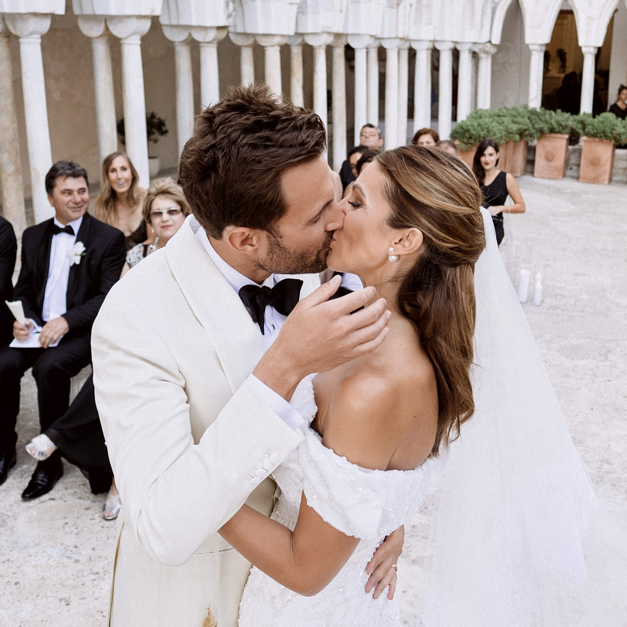 Groom in a white dinner jacket kissing the bride at the ceremony in the white-columned cloister as guests watch