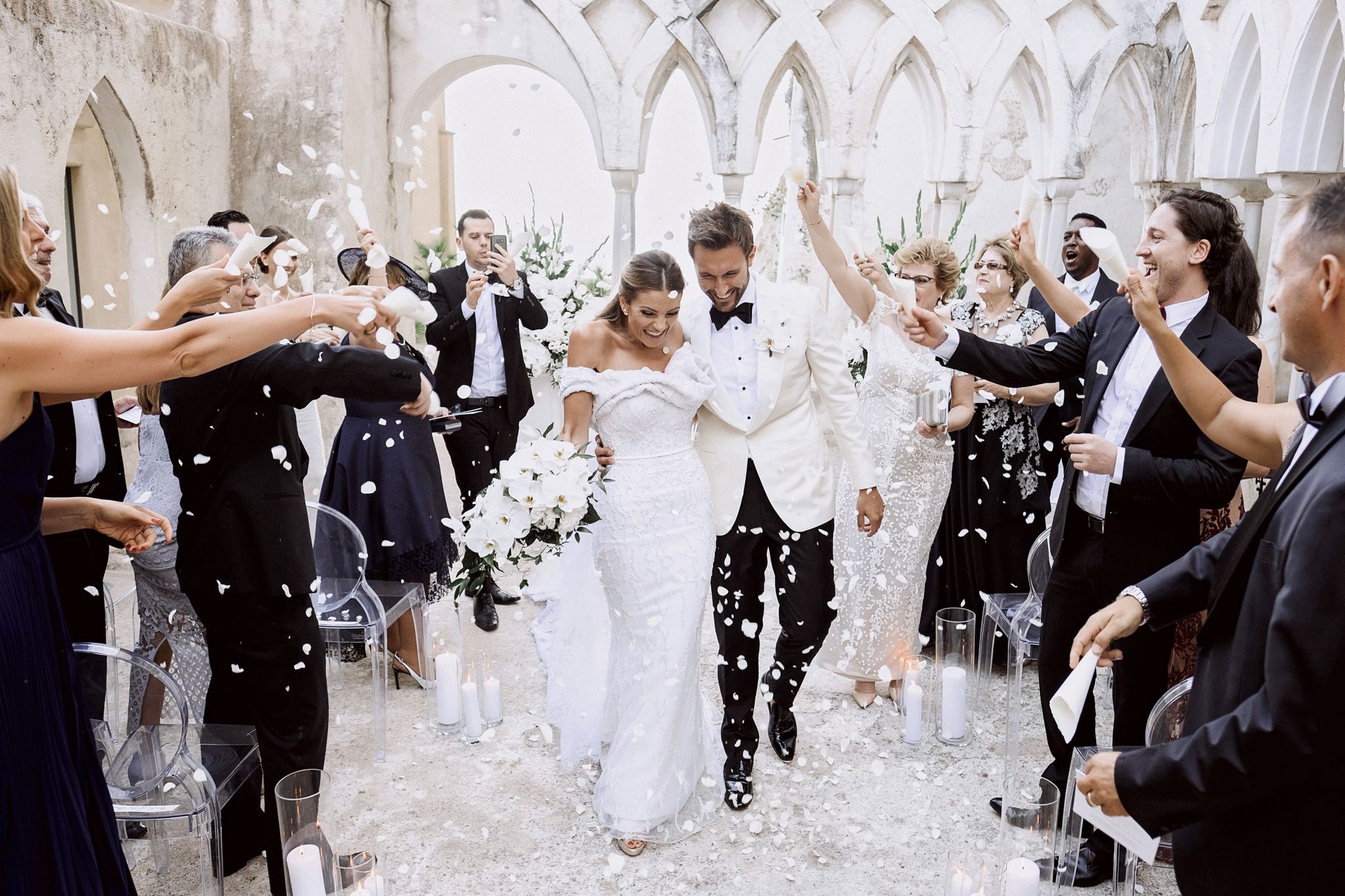 Bride and groom walking up the aisle under a shower of white petals in the Gothic-arched cloister