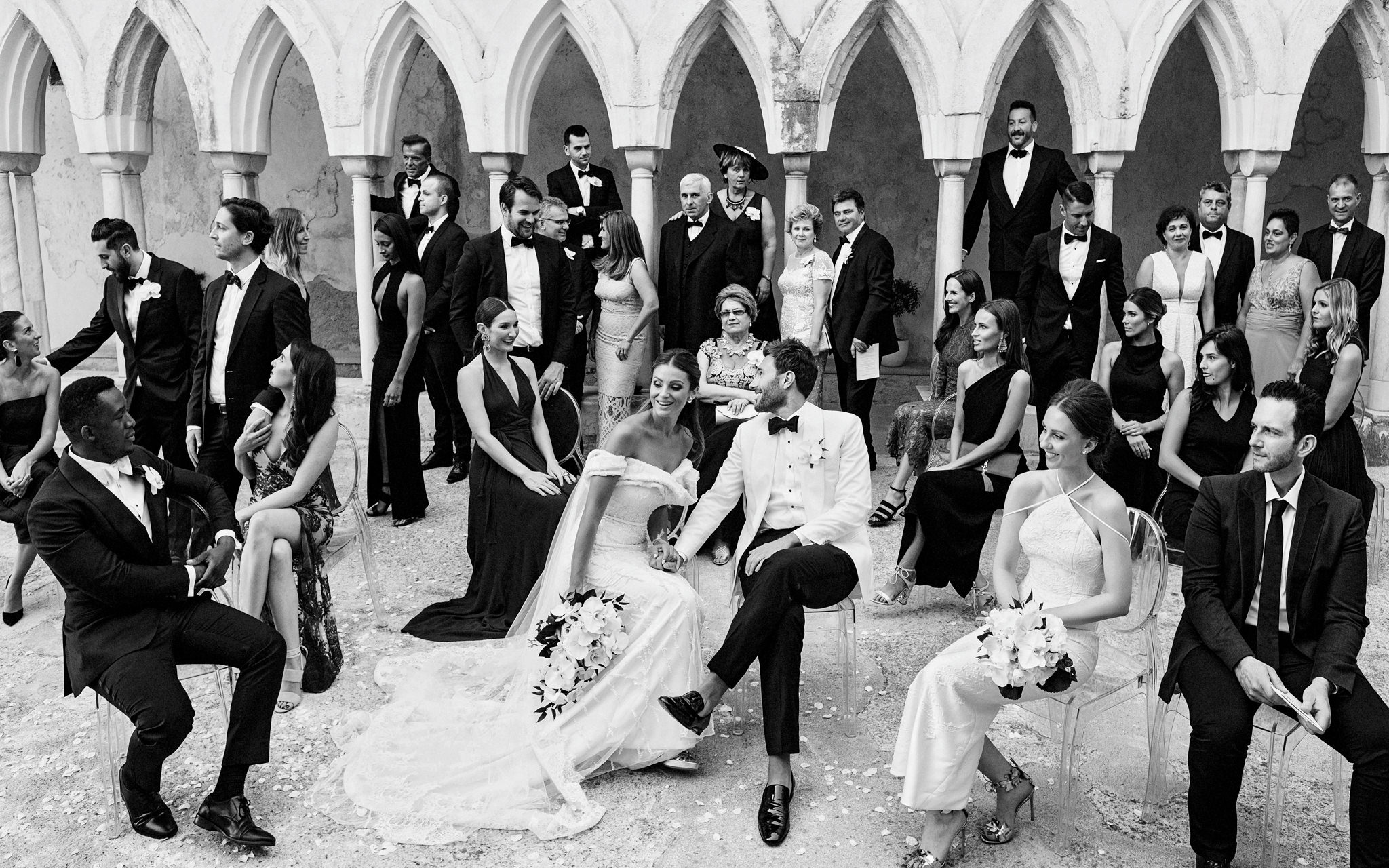 Wedding guests in black tie posed among white Gothic arches of a cloister, the bride and groom seated at centre