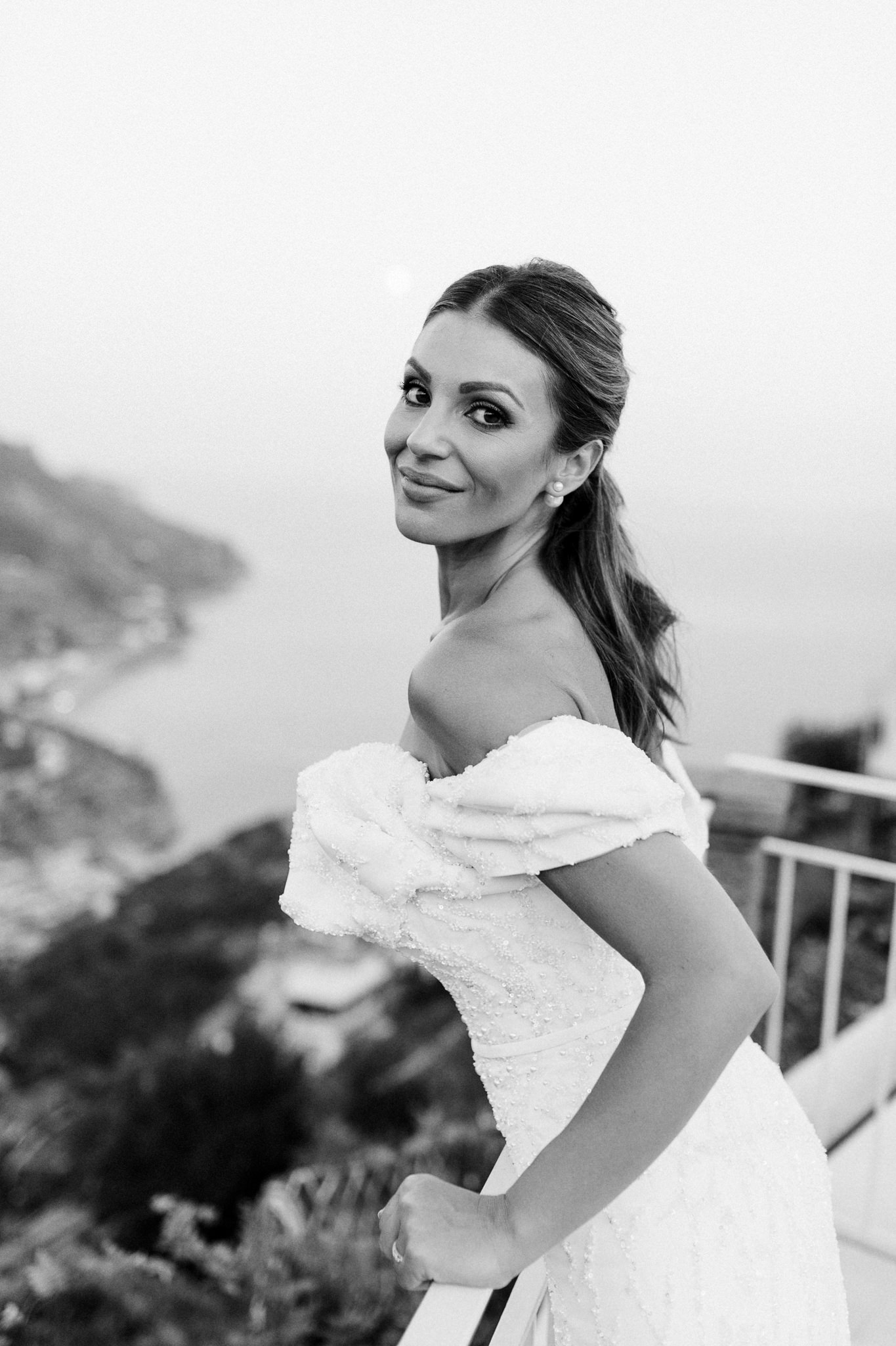 Bride in an off-the-shoulder beaded gown smiling over her shoulder on a terrace high above the coast at Ravello