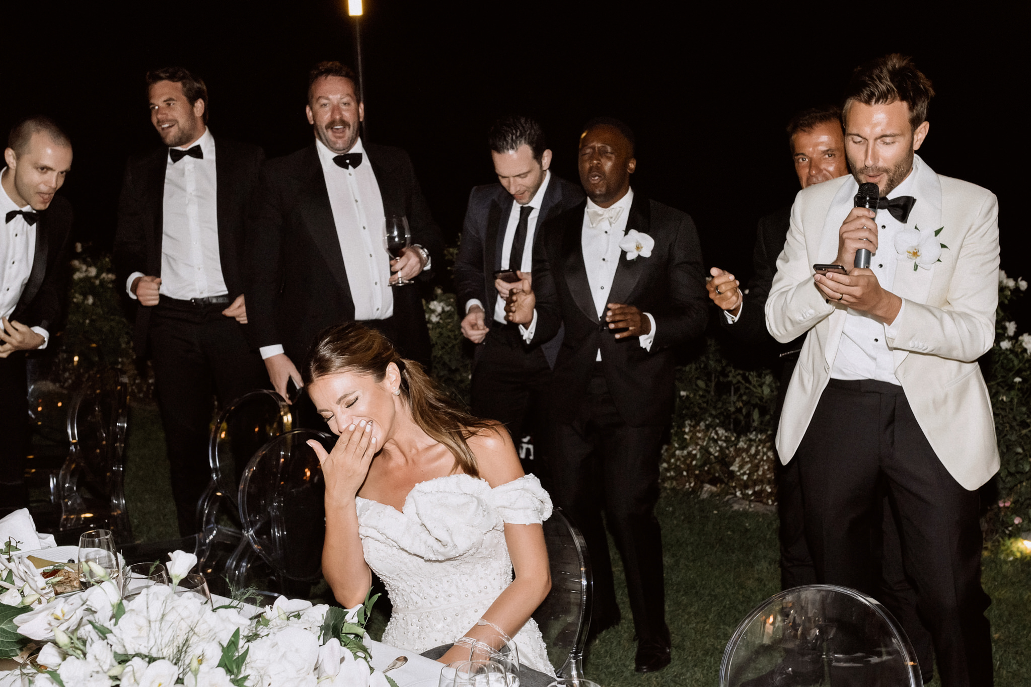 Groom serenading the bride with a microphone as friends in tuxedos sing along behind her at the dinner table