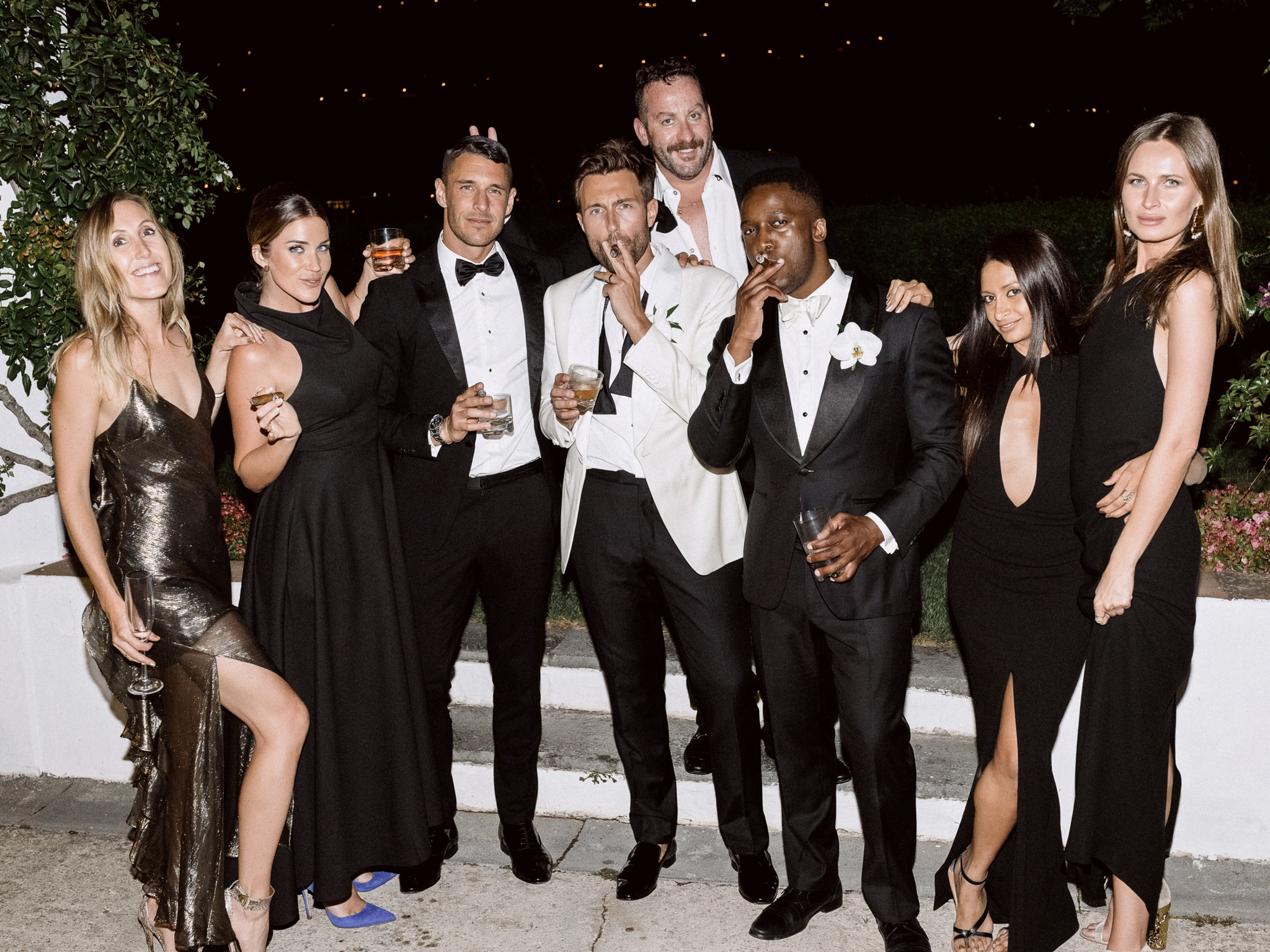 Groom and friends in black tie posing with cigars and whisky at night beside guests in black gowns